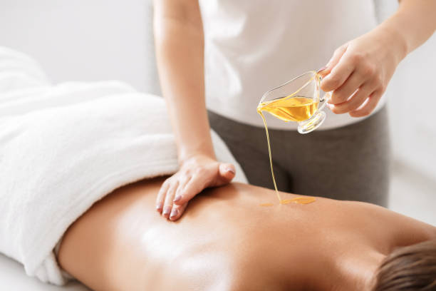 A serene spa setting where a person lies comfortably on a massage table while warm oil is gently poured onto their back. Soft lighting, candles, and calming décor create a peaceful atmosphere. A therapist uses smooth, rhythmic hand movements to spread the oil and massage the muscles, promoting relaxation and tension relief. The skin appears slightly glossy from the oil, reflecting the warm ambient light. Clean towels and natural elements like stones or plants enhance the scene. The overall mood is soothing, restorative, and tranquil, emphasizing wellness, self-care, and physical comfort in a quiet, professional environment.