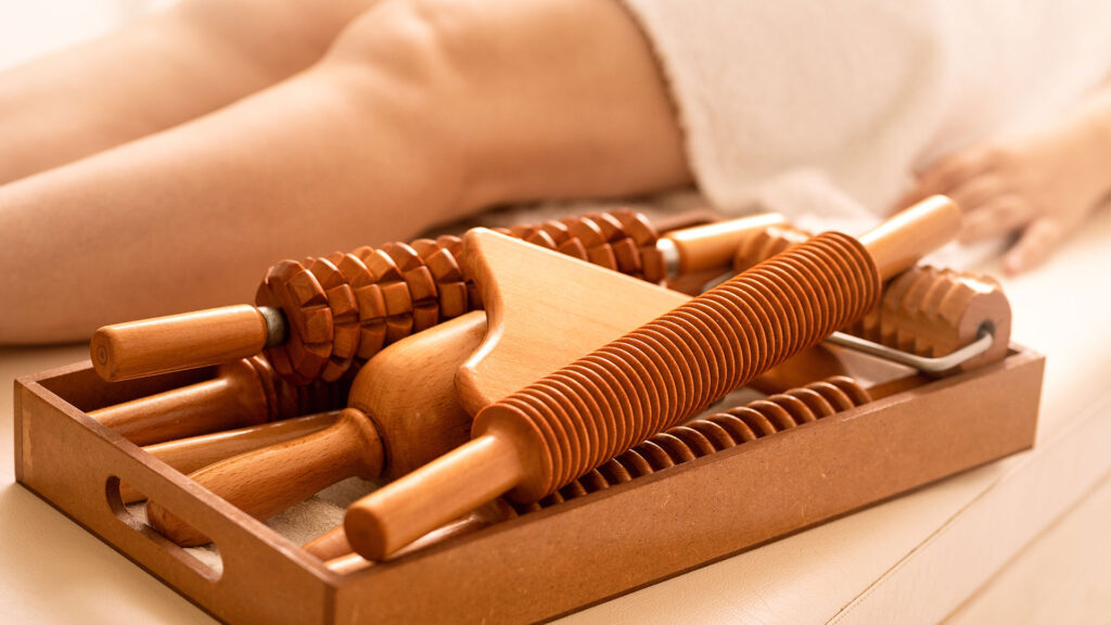 "A collection of wooden massage therapy tools, including rollers and handheld massagers, arranged in a tray beside a client lying on a massage table, representing professional massage therapy and wellness treatments.